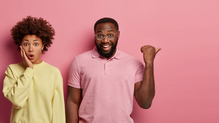 Wall Mural - Studio shot of young ethnic couple stand against pink studio background, express various emotions. Shocked Afro American woman gasps from wonder, cheerful bearded man points thumb on free space