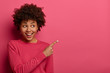Positive delighted woman with Afro haircut points index finger at upper right corner, recommends try product, wears crimson jumper, gives direction, isolated over rosy background. Check out offer