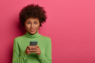 Wall Mural - Good looking woman with Afro hair types message on smartphone, browses network, looks confidently at camera, has online communication via special application, wears green poloneck. Mobile services