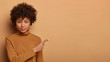 Confident serious Afro American woman persuades you going somewhere, shows direction right, points fore finger on copy space, advertises nice promo, wears turtleneck, poses over beige background.