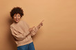 © wayhome.studio - Beautiful African American woman in beige sweater, recommends awesome product, has overjoyed face expression, stands sideways against brown background, finds perfect place to buy presents for holidays