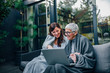 © bnenin - Happy beautiful older mother and adult daughter using laptop while sitting on wooden terrace, portrait.