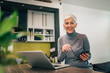© bnenin - Portrait of a happy senior woman with smart phone and laptop in the kitchen, looking at camera.