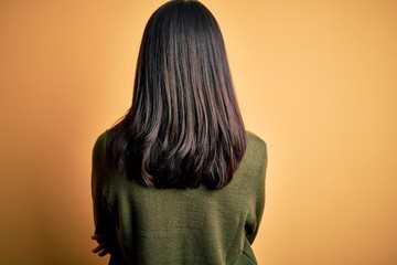 Young brunette woman with blue eyes wearing green casual sweater over yellow background standing backwards looking away with crossed arms