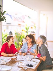 Wall Mural - Meeting of middle age women having lunch and drinking coffee. Mature friends smiling happy using smartphone at home on a sunny day