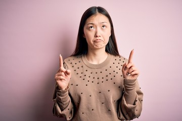 Poster - Young beautiful asian woman wearing fashion and elegant sweater over pink solated background Pointing up looking sad and upset, indicating direction with fingers, unhappy and depressed.