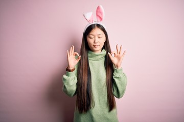 Sticker - Young asian woman wearing cute easter bunny ears over pink background relax and smiling with eyes closed doing meditation gesture with fingers. Yoga concept.