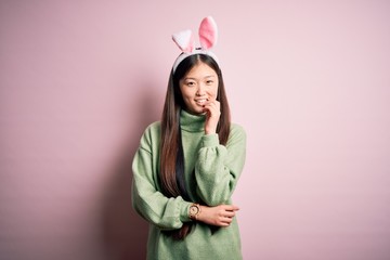 Sticker - Young asian woman wearing cute easter bunny ears over pink background looking confident at the camera with smile with crossed arms and hand raised on chin. Thinking positive.