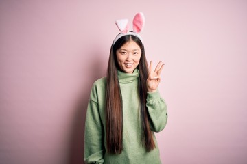 Sticker - Young asian woman wearing cute easter bunny ears over pink background showing and pointing up with fingers number three while smiling confident and happy.