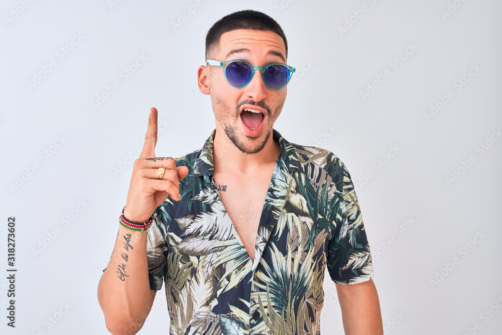Young handsome man wearing Hawaiian sumer shirt and blue sunglasses over isolated background pointing finger up with successful idea. Exited and happy. Number one.