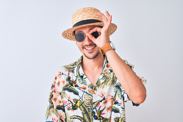 Wall Mural - Young handsome man wearing Hawaiian shirt and summer hat over isolated background doing ok gesture with hand smiling, eye looking through fingers with happy face.