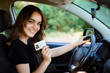 Smiling young female with pleasant appearance shows proudly her drivers license, sits in new car, being young inexperienced driver, looks with joyful expression
