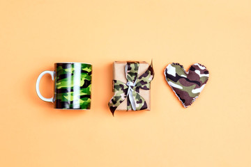 Wall Mural - Camouflage set of mug, khaki heart and gift box on yellow  background. Top down composition.