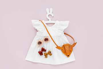 White dress  with kids handbag and sunglasses. Set of  baby clothes and accessories for spring or summer on pale pink background. Fashion childs outfit. Flat lay, top view