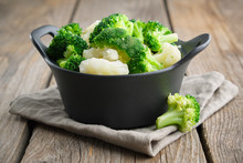 Black Saucepan With Cooked Broccoli And Cauliflower On Wooden Table.