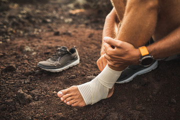 Wall Mural - Man bandaging injured ankle. Injury leg while running outdoors. First aid for sprained ligament or tendon. Close-up on dark background.