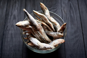 Frozen sprat herring fish in a glass bowl on a wooden dark background.