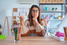 Young Beautiful Teacher Woman Wearing Sweater And Glasses Sitting On Desk At Kindergarten Asking To Be Quiet With Finger On Lips Pointing With Hand To The Side. Silence And Secret Concept.