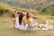 The company of cheerful female friends in white dresses enjoys a view of the green hills, relaxing on a picnic.