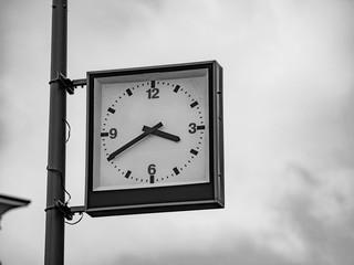 Street clock with a traditional dial and hands arrows for hours and minutes. An rectangular boxed analog clock on a city lighting pole shows the exact time 3:40 pm. Black and white shot