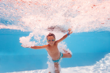 Happy little boy splash into the pool after jump making many bubbles, smiling and wearing swimming googles