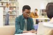 © Seventyfour - Portrait of African teenage boy studying in college library, using laptop with other students in background, copy space