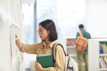 Wall Mural - Side view portrait of female Asian student wearing glasses choosing books on shelves in college library, copy space