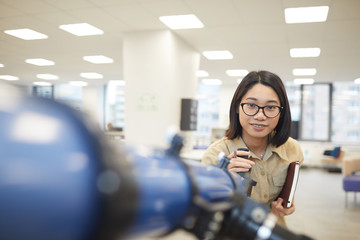 Wall Mural - Portrait of cheerful Asian girl looking at camera while posing by telescope in astronomy class or college library, copy space