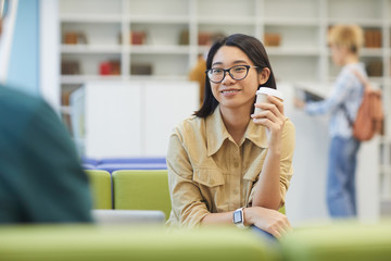 Wall Mural - Portrait of smiling Asian girl enjoying coffee while studying in college library, copy space