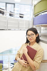 Wall Mural - Waist u portrait of young Asian woman smiling at camera while standing by spiral staircase in modern college interior, copy space