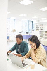 Wall Mural - High angle portrait of two students studying in college library, focus on Asian young woman using laptop in foreground, copy space