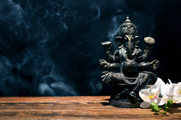 Hindu god Ganesh on black background. Statue on wooden table with a smoke of incense. Copy space.