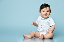 Little Kid In White Bodysuit As Vest With Bow-tie, Barefoot. He Smiling, Sitting On The Floor Against Blue Background. Close Up