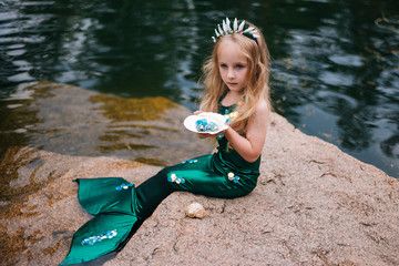 a little girl with white hair with a mermaid tail and shells is sitting on the shore. a little merma