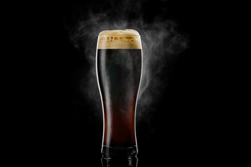 Cold pilsner beer glass with black porter beer inside covered with drops and froth evaporating on black background.