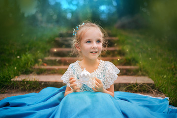 Little girl with white hair in a blue retro dress holds a small carriage in her hands. Princess from a fairy tale sitting on the stairs