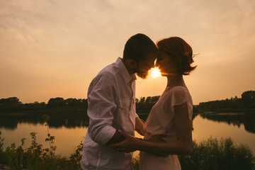 Wall Mural - Couple standing on a river shore, kissing while sun is setting