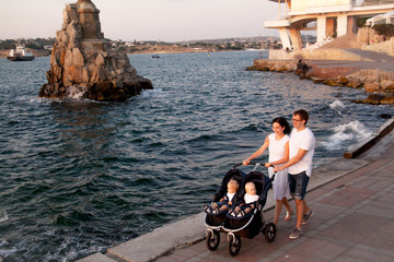 Wall Mural - Happy family walking with twins buggy on promenade against the sea.