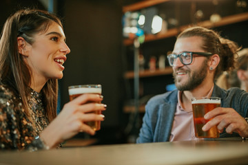 Wall Mural - Young handsome caucasian bearded man with eyeglasses leaning on bar counter in pub, drinking beer and chatting with friend.