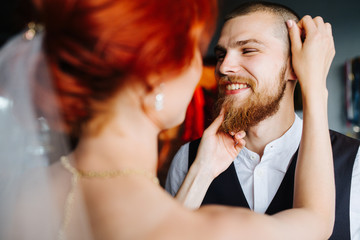 Beautiful bride and groom together in the bright room before the marriage ceremony. She's patting and grooming her future husband. He giggles, feeling a little anxious.