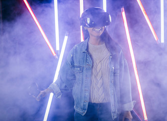 Mod curly dark haired girl dressed in blue denim jacket uses the virtual reality glasses on her head and gloves in hands in the dark studio with neon light and smoke fog