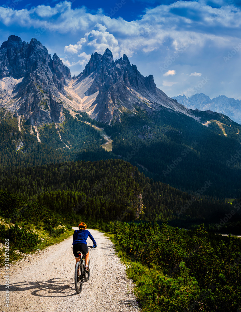 Tourist cycling in Cortina d'Ampezzo, stunning rocky mountains on the background. Woman riding MTB enduro flow trail. South Tyrol province of Italy, Dolomites. - obrazy, fototapety, plakaty 