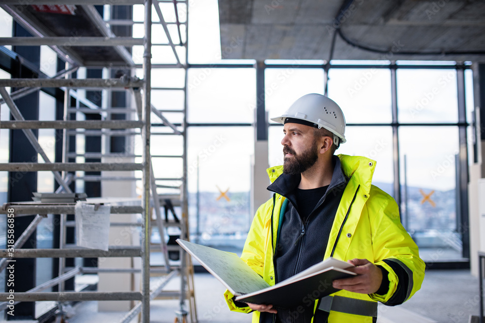 Man engineer standing on construction site, holding blueprints. - obrazy, fototapety, plakaty 