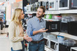 © LIGHTFIELD STUDIOS - Smiling consultant pointing with hand at microwave and talking with woman in home appliance store