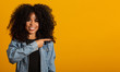 © Brastock Images - Cheerful Afro woman points away on copy space, discusses amazing promo, gives way or direction, wears yellow warm sweater, has pleasant smile, feels optimistic, isolated over yellow background.