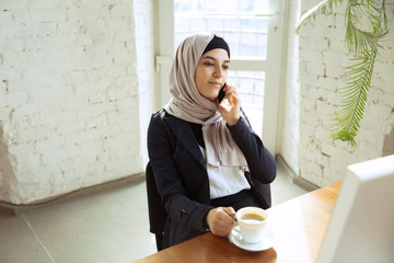 Wall Mural - Talking on phone while drinking coffee. Portrait of a beautiful arabian businesswoman wearing hijab while working at openspace or office. Concept of occupation, leadership, success, modern solution.