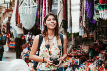 Hispanic Woman Backpacker Holding A Camera In A Traditional Mexican Market In Mexico, Vacations
