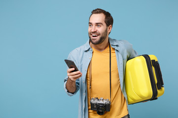 Wall Mural - Funny traveler tourist man in yellow clothes with photo camera isolated on blue background. Male passenger traveling abroad on weekends. Air flight journey concept. Hold suitcase, using mobile phone.