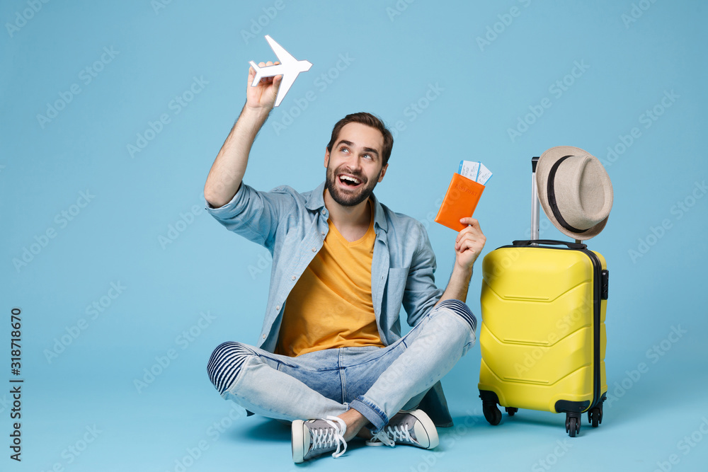 Funny traveler tourist man in yellow clothes isolated on blue background. Passenger traveling abroad on weekend. Air flight journey Sit near suitcase hold passport boarding pass tickets air plane. - obrazy, fototapety, plakaty 