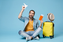 Funny Traveler Tourist Man In Yellow Clothes Isolated On Blue Background. Passenger Traveling Abroad On Weekend. Air Flight Journey Sit Near Suitcase Hold Passport Boarding Pass Tickets Air Plane.